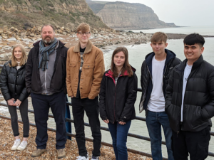 A group of students pose for a photo with a staff member in front cliffes by the sea.